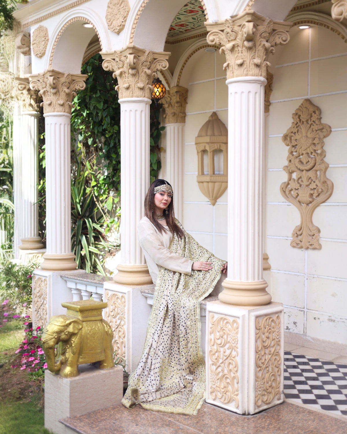 A woman wearing a traditional Indian salwar kameez outfit with a matching printed saree stands on an ornate outdoor veranda with cream pillars and decorative carvings.
