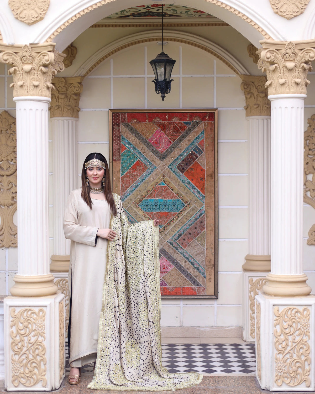 A woman in a cream-colored traditional outfit stands in an ornate architectural setting with columns, holding a long patterned quilt or shawl. Behind her is a large framed multicolor geometric textile hanging on the wall.