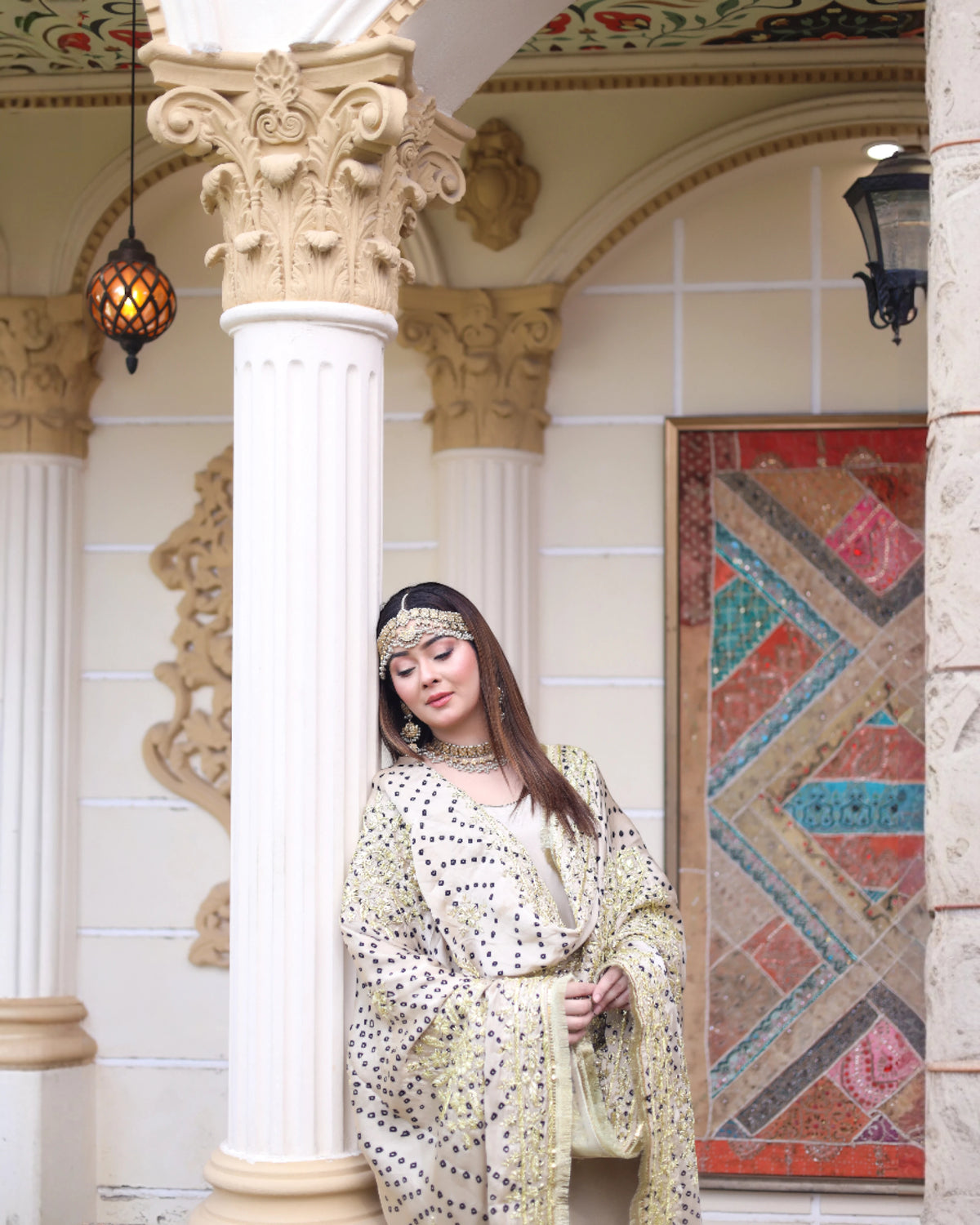 A woman wearing a cream and gold embroidered traditional outfit with a matching headpiece stands in front of an ornate beige column in a decorated architectural setting.