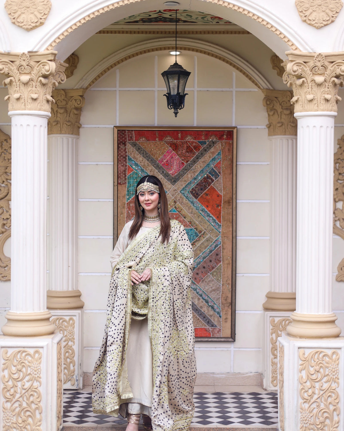 A woman stands in an ornate architectural doorway wearing a traditional outfit with a patterned shawl. The setting features cream-colored columns, a hanging lantern, and a colorful geometric tapestry on the wall behind her.