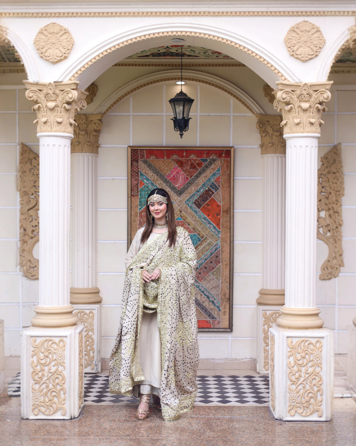 A woman stands under an ornate white archway with carved columns, wearing a flowing beige and green patterned outer garment over a cream dress. A colorful patterned tapestry hangs behind her, and a black hanging lantern is suspended above.

