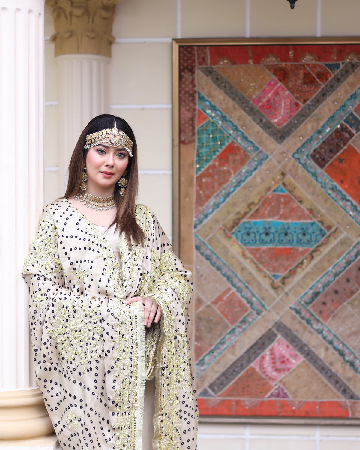 A woman models a traditional embroidered white and beige kaftan dress with black dotted patterns, styled with gold jewelry, standing in front of a decorative geometric wall art piece.