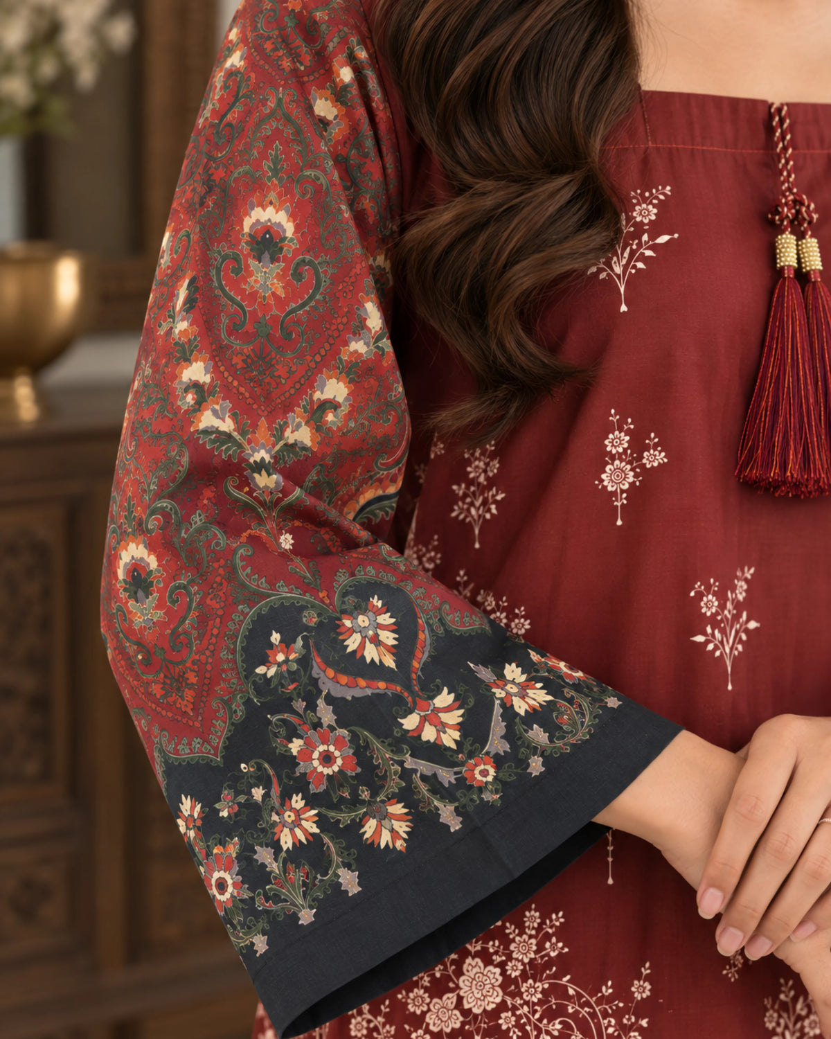A woman wearing a deep red kurta with beige floral prints, featuring a square neckline, a decorative tassel at the center, and patterned sleeves with dark navy borders.