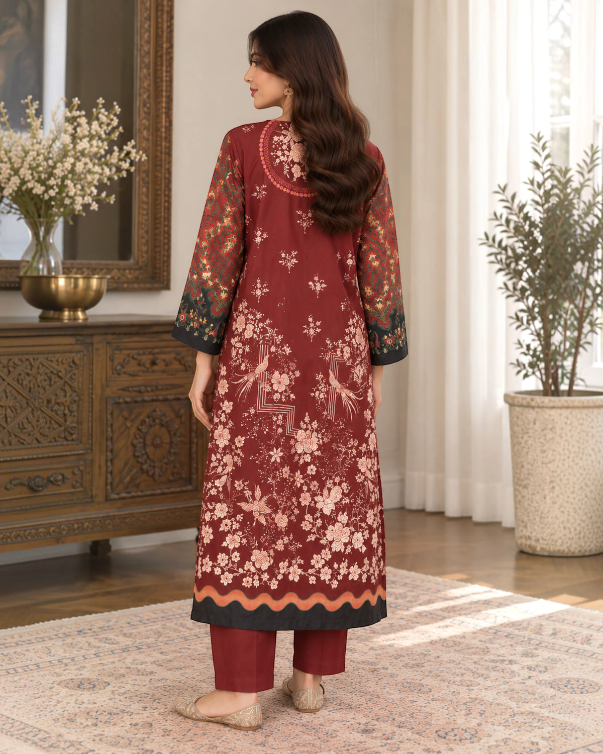 A woman stands with her back to the camera wearing a long burgundy embroidered kurta with matching trousers in a decorated room with plants and a wooden cabinet.