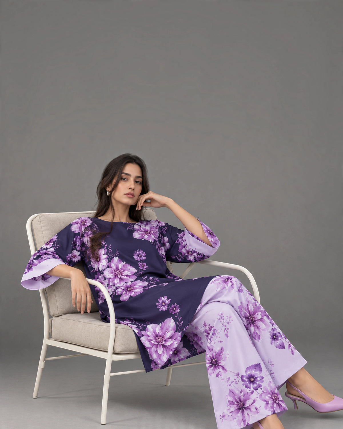 A woman models a matching floral printed kurta and trouser set while sitting in a modern white armchair against a solid gray studio background.